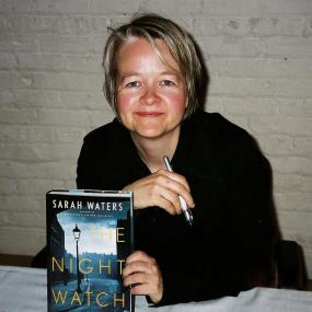 Colour photograph of Sarah Waters displayng a copy of her book "The Night Watch", 2006. She leans forward, with short blonde-streaked hair, blue eyes, and a smile, wearing a black shirt and holding a pen.