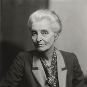 Black and white photograph of Beatrice Webb, seated with one arm resting on the arm of her chair. She is wearing a dark jacket with a light collar, and a polka-dotted tie. Her white hair is pulled back.