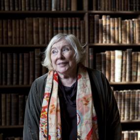 Colour photo of Fay Weldon standing in a library at an event during the Oxford Literary Festival, 3 April 2011. She wears a grey top and a
            colourful patterened scarf. 
