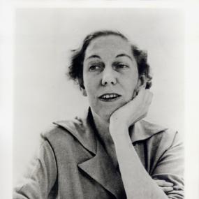 Black and white photograph of Eudora Welty. She sits looking up with her lips parted, her chin resting on her left hand and her other arm across her body. She is wearing a shirt with a broad collar.