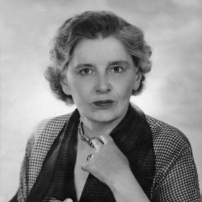 Black and white, head-and-shoulders photograph of Rebecca West looking straight forward, wearing a checked jacket with dark cuffs and collar and white flowers pinned at the neckline. With one hand she touches a chunky metal necklace. Her wavy, greying hair, jaw-length, is pushed behind her ears.