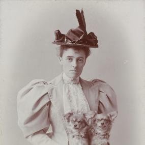 Monochrome cabinet photograph of Edith Wharton, taken by E. F. Cooper, at Newport, Rhode Island. Courtesy of the Beinecke Rare Book and Manuscript Library, Yale University. She is seated looking at the camera, in a jacket with leg-of-mutton sleeves and tight bodice, with a fall of lace in front, and a small hat with two feathers sticking up. She has two little dogs on her lap.