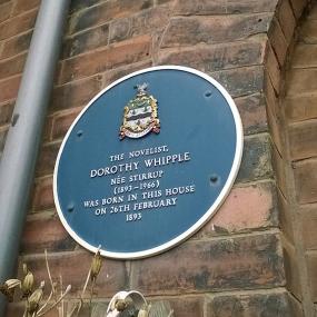 Colour photograph of a blue plaque on the brick wall of Dorothy Whipple's birthplace in Blackburn, Lancs. The plaque has a coat of arms at the top, and reads "The novelist Dorothy Whipple (née Stirrup) (1893-1966) was born in this house on 26th February 1893."
