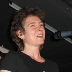 Photograph of Janette Winterson taken in Warsaw, Poland, on 16 February 2005 by Mariusz Kubik. She has her lips parted, and is apparently speaking into a microphone . She has short, brown,curly hair and wear a black t-shirt with a seahorse logo.