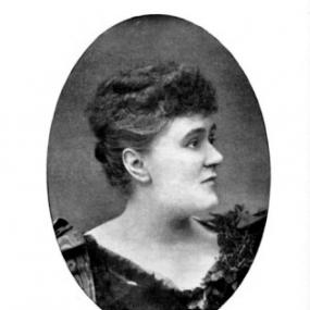 Black and white, oval photograph of John Strange Winter. Her face is in profile, her body turned a little to the right. Her curly hair is styled in a fringe in front and a bun behind.  She is wearing a black dress with a tulle-like fabric at the neck. Her signature is reproduced below: "Ever yours, H. E. V. Stannard".