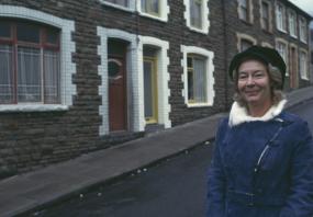 Photo of Nina Bawden in front of a row of small houses with stone facades, wearing a blue coat and a black hat, 1974. 