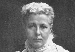 Photograph of Annie Besant looking directly at the camera with her right hand over her chest. She has a serious expression and is wearing a white high frill-collared dress with possibly a shawl over it. She has two rings on her visible right hand. She has blonde hair and it is in an updo.