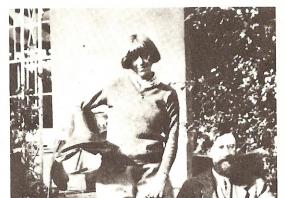 Black and white photo of Dora Carrington standing beside a seated Lytton Strachey in a garden. She is wearing jodhpurs, riding boots, and a sweater over a shirt. Her hair is bobbed and she holds a horse's saddle with the left stirrup hanging down.