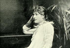 Black and white photograph of Harriett Jay, seated in a chair and leaning her elbow on a desk, with a potted plant behind. She is wearing a long, simple, white dress with lace trim at the sleeves and throat, and a silver bracelet and ring. Her light hair is pulled back, with short curls in front.