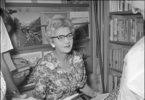 Black-and-white photo of Norah Lofts sitting at a desk for a book signing. Behind her are stacks of bookstore shelves. She has cat-eye glasses
            and wears a floral dress. People are gathered around her holding her books out to be signed. 