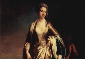 Photo of a full-length painting of Lady Mary Wortley Montagu by Jonathan Richardson recording her time in Turkey. She stands outdoors, with famous buildings of Constantinople (Istanbul) visible beyond. She is dressed in gold: a caftan whose deep V-neck and open skirt reveal a lace shift and long harem drawers. She also wears a gold cap which she calls a talpock on her dark hair, a blue cloak edged with ermine, pearl drop earrings, and pointed slippers. The presence of the young Black boy behind her is start