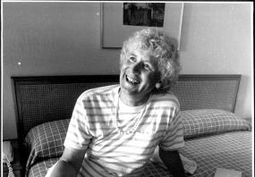 Black-and-white photo of Jan Morris seated on what looks like a hotel bed, smiling. 