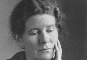 Black and white photograph of E. J. Scovell, shown from the shoulders up. She is wearing a collared corduroy jacket with buttons up the front, and her curly hair is brushed back from a centre parting. Her head is resting on her hand. She looks thoughtful.