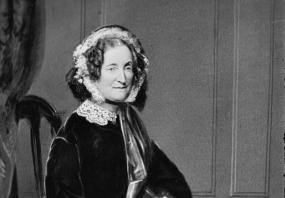 Full-length photograph of Lydia Howard Sigourney by Matthew Brady. She is seated on a wooden chair with wooden panelling behind her, wearing a dark jacket and hood with lace bonnet and collar, and a full, boldly checked skirt. She holds a handkerchief in her hands.