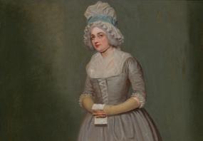 Photo of a full-length painting of Leah Sumbel by Samuel de Wilde, performing (under her original name, Mary Wells) as Anne Lovely in Susannah Centlivre's "A Bold Stroke for a Wife". She stands on stage, wearing a long, plain grey dress laced up the bodice, its square neckline filled with a fichu, Her hair is powdered under a tall mob cap with a blue ribbon, and she holds a paper in her hands.