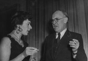 Black-and-white photo of Edith Templeton and V S Pritchett smoking cigarettes as they chat at a party celebrating the 25th anniversary of the
            Book Society, Knightsbridge, London, 1946.