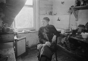 Black-and-white photo of Anna Wickham in her cluttered kitchen, originally published to accompany "Anna Wickham: A Poetess Landlady" in
            Picture Post, 1946. Looking off-camera, she is seated on a chair and smoking a cigarette. 