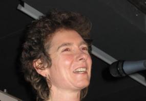 Photograph of Janette Winterson taken in Warsaw, Poland, on 16 February 2005 by Mariusz Kubik. She has her lips parted, and is apparently speaking into a microphone . She has short, brown,curly hair and wear a black t-shirt with a seahorse logo.