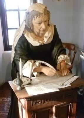Colour photograph of a mannequin representing Anne Halkett, sitting at a desk writing, dressed in black with white cap, collar, and cuffs. The picture was taken at the heritage centre (since 2015 struggling for financial support) in Abbot House, Dunfermline, the oldest building in the town where Halkett spent her last three decades.